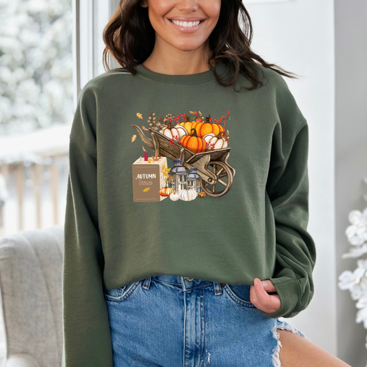 Person wearing a green sweatshirt with a fall-themed design  featuring a graphic design of a rustic wooden wheelbarrow overflowing with various orange and white pumpkins, berries, and autumn leaves. A small box with "Autumn Stories" written on it sits next to two lit lanterns.