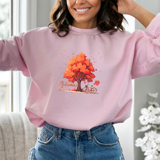Person wearing a pink sweatshirt with an autumn tree designfeaturing a graphic design of a large autumn tree with vibrant orange and red leaves. A red bicycle with a basket and heart-shaped balloons is parked beside the tree. The word "Autumn" is written in a flowing script font to the left, and numerous fall leaves are scattered around the scene, some falling from the tree.