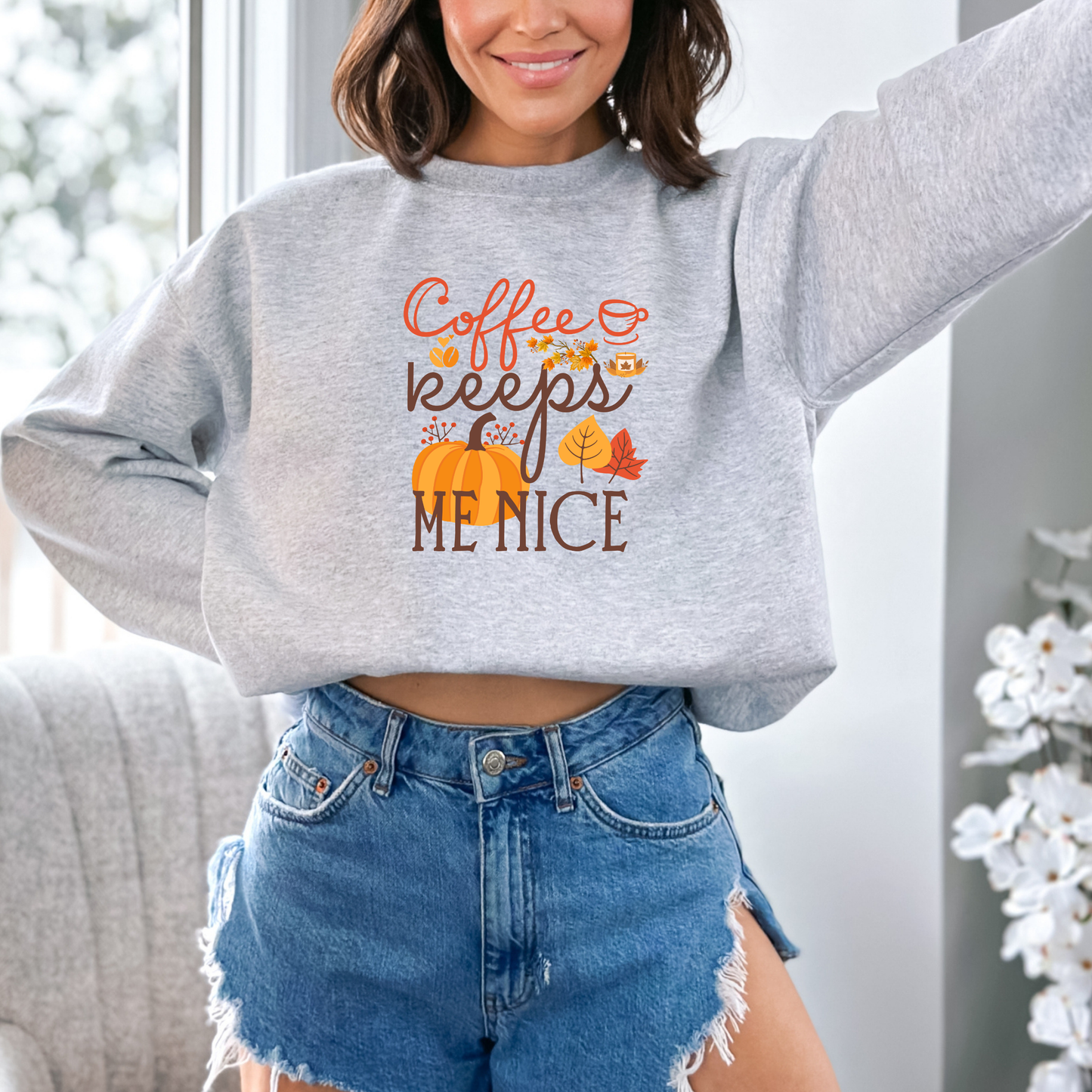 A girl wearing a grey sweat shirt featuring a graphic design with the text "Coffee keeps ME NICE" in a mix of orange cursive and brown block fonts. The design is decorated with a large orange pumpkin, autumn leaves, red berries, coffee beans, a lit candle, and a small coffee cup.