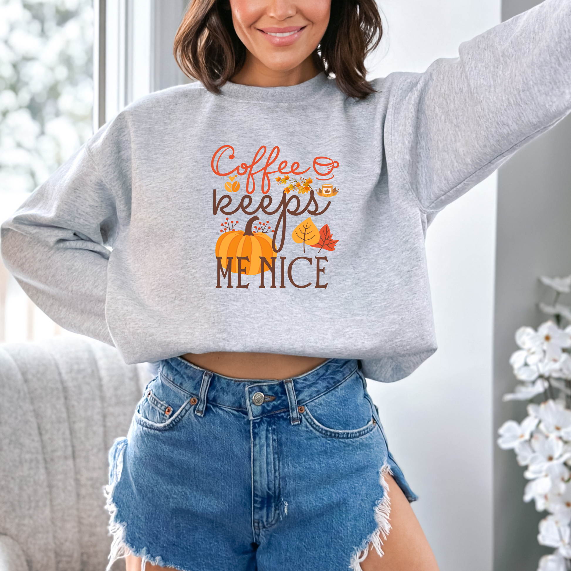 A girl wearing a grey sweat shirt featuring a graphic design with the text "Coffee keeps ME NICE" in a mix of orange cursive and brown block fonts. The design is decorated with a large orange pumpkin, autumn leaves, red berries, coffee beans, a lit candle, and a small coffee cup.