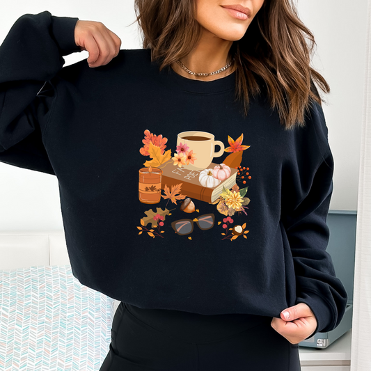 Person wearing a black sweatshirt with a graphic design featuring a graphic design of various cozy autumn items. A steaming mug of tea with a tea bag rests on top of a closed brown book. Next to it is a lit orange candle. The scene is decorated with scattered autumn leaves, red berries, chestnuts, and a pair of eyeglasses.