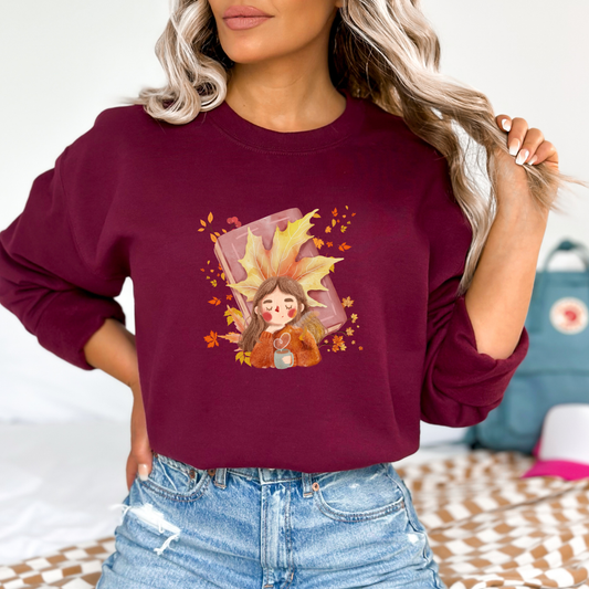 Person wearing a maroon sweatshirt featuring a graphic design of a cute cartoon girl with brown hair, wearing an orange sweater and holding a light blue mug with a heart-shaped steam. She is depicted resting or dreaming, with a large yellow maple leaf behind her head and a closed pink book behind the leaf. The design is surrounded by falling autumn leaves and berries.