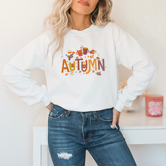 Person wearing a white sweatshirt with 'AUTUMN' and autumn-themed graphics featuring a graphic design with the word "AUTUMN" in large, decorative letters filled with various patterns and small illustrations. The design also includes a small owl, two flying birds, hot drinks in mugs, pumpkins, acorns, and scattered leaves and socks.