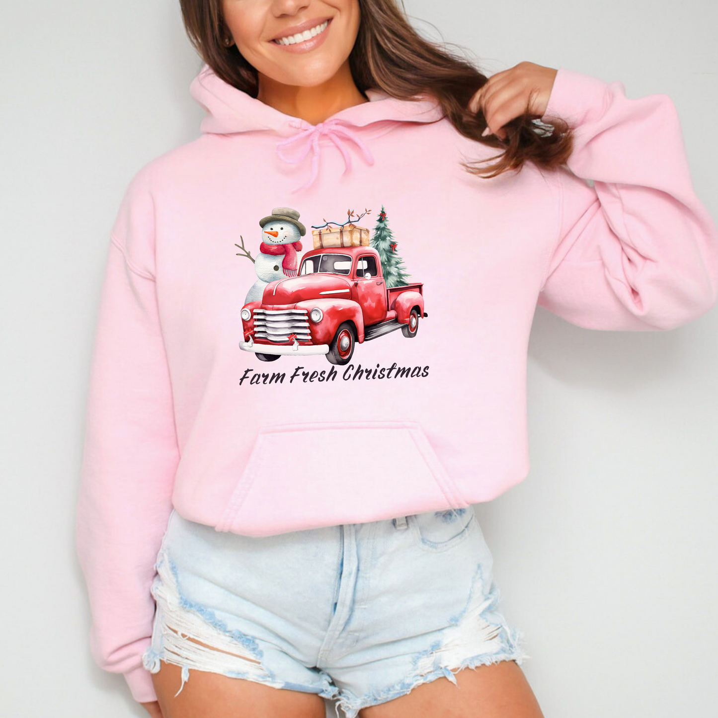 Person wearing a pink hoodie with a Christmas design on a plain background featuring a graphic design of a vintage red pickup truck with a large snowman and a small Christmas tree in the back. The truck has a suitcase on top of the bed. Below the truck, the words "Farm Fresh Christmas" are written in a festive, handwritten font.