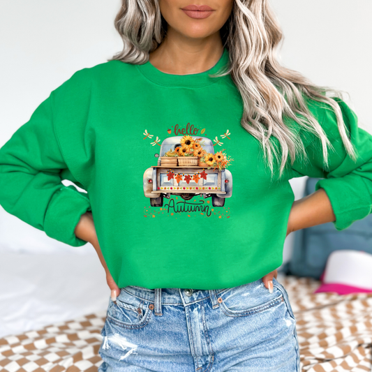 Person wearing a green sweatshirt with an autumn design featuring afeaturing a graphic design of the back of a vintage light blue pickup truck. The truck bed is filled with crates of sunflowers, pumpkins, and other fall foliage. A garland of colorful autumn leaves hangs from the tailgate. Above the truck, the word "hello" is in a script font, and below the truck, "Autumn" is in a matching script font, surrounded by swirling fall leaves and dragonflies.