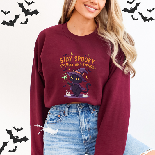Person wearing a maroon sweatshirt with Halloween design and text, surrounded Light gray long sleeve shirt featuring a graphic design with the text "STAY SPOOKY" in a dripping orange font. Below the text, a cute black cat is dressed as a witch, wearing a purple hat and cape and holding a glowing green wand. The cat is surrounded by stars, moons, and bats, and the words "FELINES AND FIENDS" are between the cat and the main text.