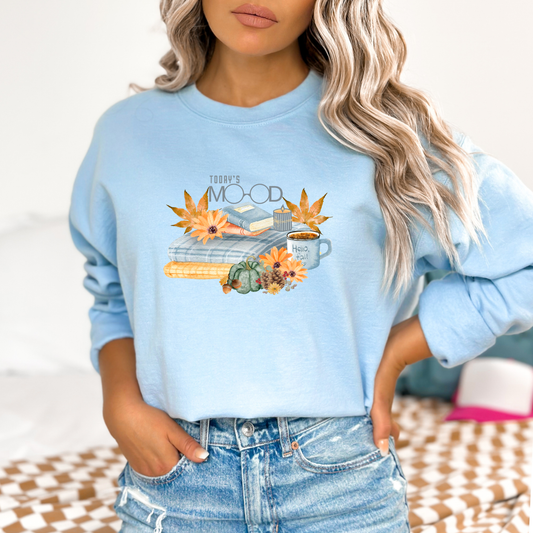 Light blue sweatshirt with autumn-themed design eaturing a graphic design with the text "TODAY'S MOOD" in gray above an illustration of cozy fall items. The items include a stack of plaid and textured blankets, two books, a lit candle, a mug with "Hello, fall" written on it, orange and green pumpkins, acorns, pinecones, and various fall leaves and flowers in shades of orange and brown worn by a person.