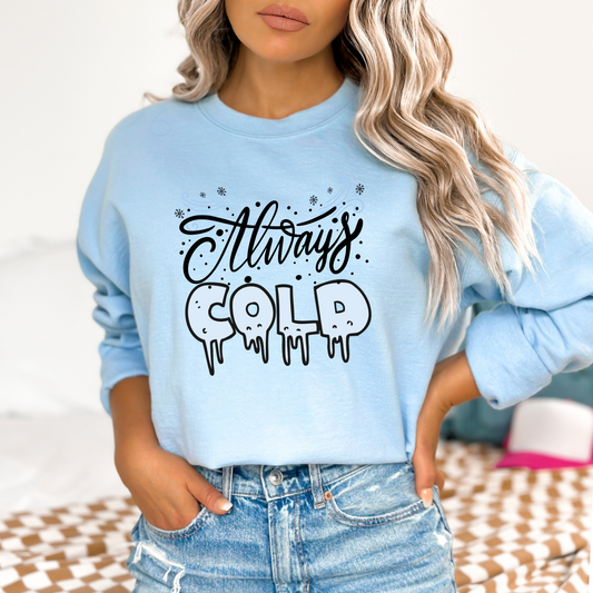 Light blue sweatshirt with 'Always Cold' text worn by a person, on a neutral background featuring a graphic design with the words "Always" in a black, handwritten cursive font and "COLD" in large, light blue, dripping, icicle-like letters. The design is decorated with small black dots and light blue swirls and snowflakes.