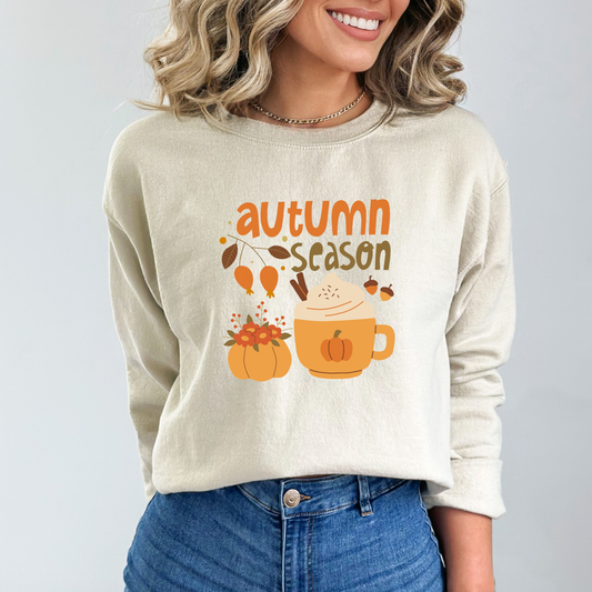 Person wearing a beige sweatshirt featuring a graphic design with the words "autumn season" in an orange, stylized font. The design includes a large, cozy mug of a hot drink topped with whipped cream and cinnamon sticks, a pumpkin-shaped vase with flowers, and two acorns.