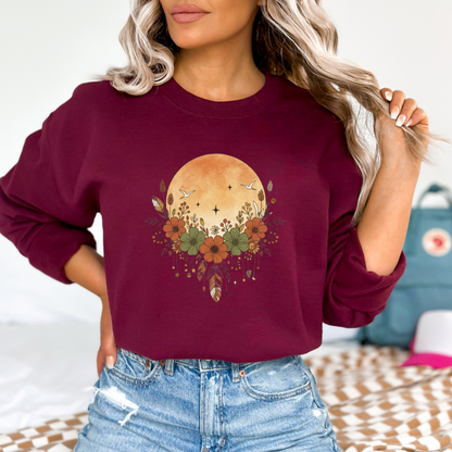 sweatshirt featuring a boho-style graphic with a large, orange, textured full moon in the background. In the foreground is a decorative arrangement of rustic flowers in shades of burnt orange and olive green, surrounded by wheat stalks, berries, and feathers hanging below. Two white birds fly across the moon.
