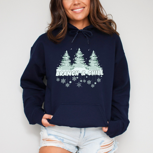 Navy blue hoodie with 'Branch & Chill' design worn by a person against a light background hooded sweatshirt featuring a graphic design with the phrase "Branch & Chill" in a thick, rounded font. Above the text is a stylized winter scene with three snow-covered pine trees and falling snow. The entire design is accented with delicate snowflake illustrations below the text.