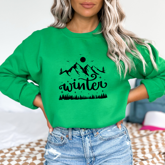 Person wearing a green sweatshirt with a mountain and 'winter' design featuring a black graphic design with the cursive word "winter" prominently displayed in the center. Above the text are three stylized silhouette mountains, a full moon, and three flying birds. Below the word "winter" is a silhouette of a row of pine trees. The design includes scattered dots and starbursts to suggest snow or a starry night.