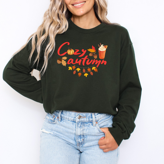 Person wearing a green sweatshirt with 'Cozy Autumn' text and fall-themed graphics on a white background featuring a graphic design with the words "Cozy autumn" in a stylized red cursive font. The letter 'o' in "Cozy" is replaced by a round, textured pumpkin. The design is decorated with a variety of fall illustrations, including acorns, autumn leaves, and a hot beverage in a transparent cup topped with whipped cream and cinnamon sticks. A garland of colorful leaves hangs below the text.