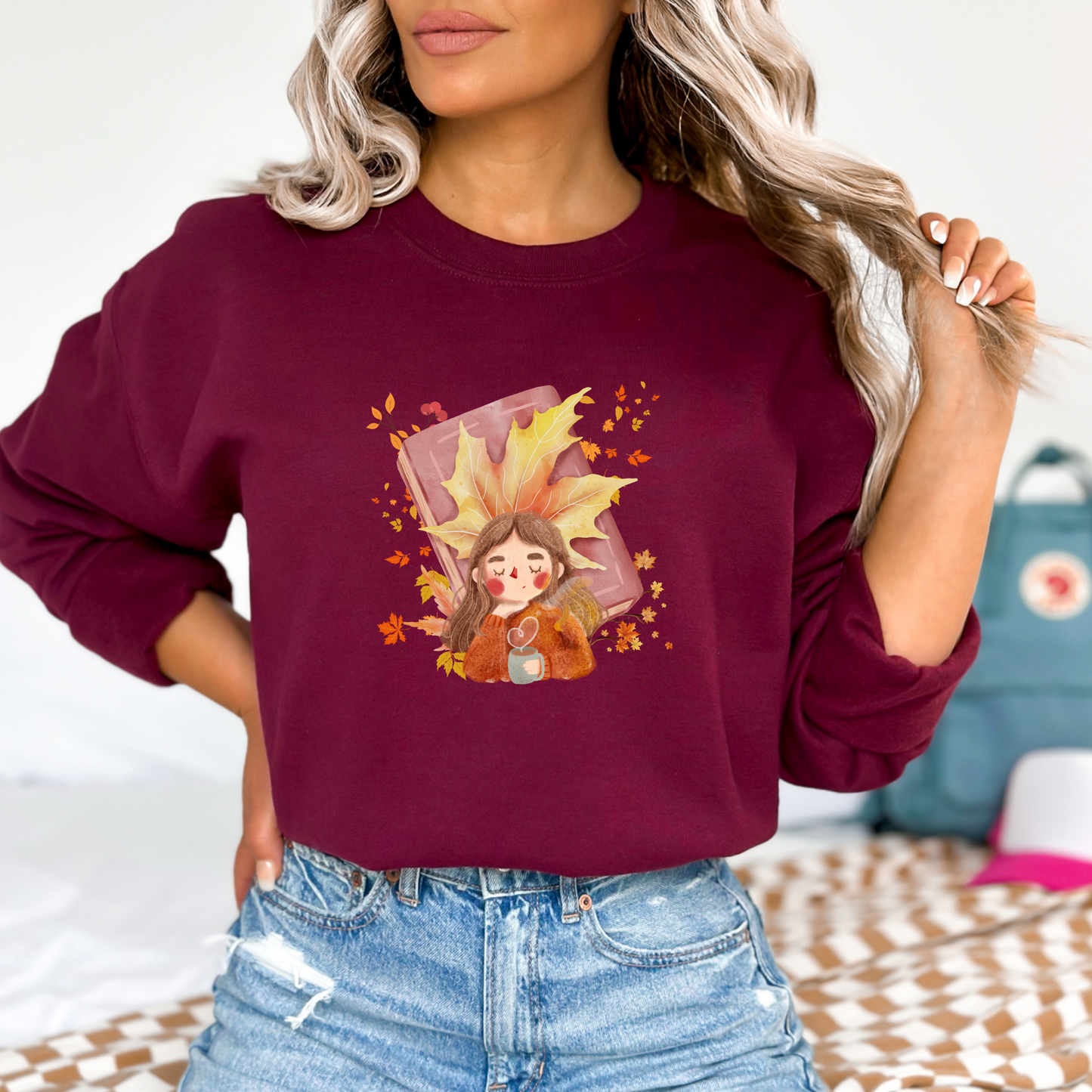 Person wearing a maroon sweatshirt featuring a graphic design of a cute cartoon girl with brown hair, wearing an orange sweater and holding a light blue mug with a heart-shaped steam. She is depicted resting or dreaming, with a large yellow maple leaf behind her head and a closed pink book behind the leaf. The design is surrounded by falling autumn leaves and berries.