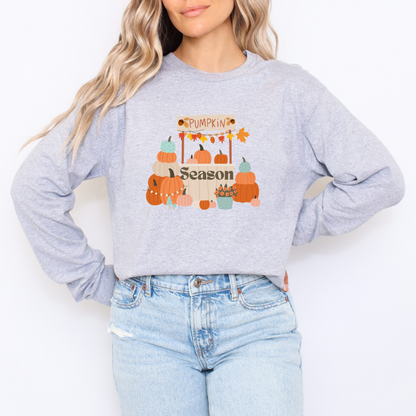 Person wearing a light gray sweatshirt with 'Pumpkin Season' design and light blue jeans on a white background featuring a graphic design of a pumpkin patch stand. The stand has a sign that reads "PUMPKIN" decorated with leaves and acorns. Below the sign, a variety of pumpkins in different colors (orange, white, light blue, and pink) are stacked and arranged around the stand. The word "Season" is on the front of the stand, and a blue pot of orange flowers sits next to it.
