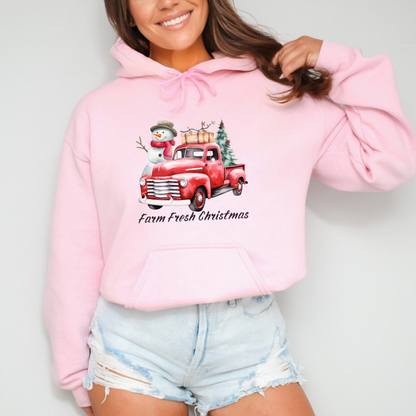 Person wearing a pink hoodie with a Christmas design on a plain background  featuring a graphic design of a vintage red pickup truck with a large snowman and a small Christmas tree in the back. The truck has a suitcase on top of the bed. Below the truck, the words "Farm Fresh Christmas" are written in a festive, handwritten font.
