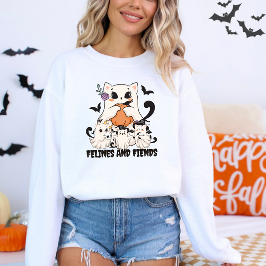 Person wearing a white sweatshirt with with a graphic of a large, cute cat dressed as a ghost, holding a pumpkin. Below it, three smaller cats in ghost costumes pose with a skull and a witch hat. The words "FELINES AND FIENDS" are at the bottom with Halloween decorations in the background.