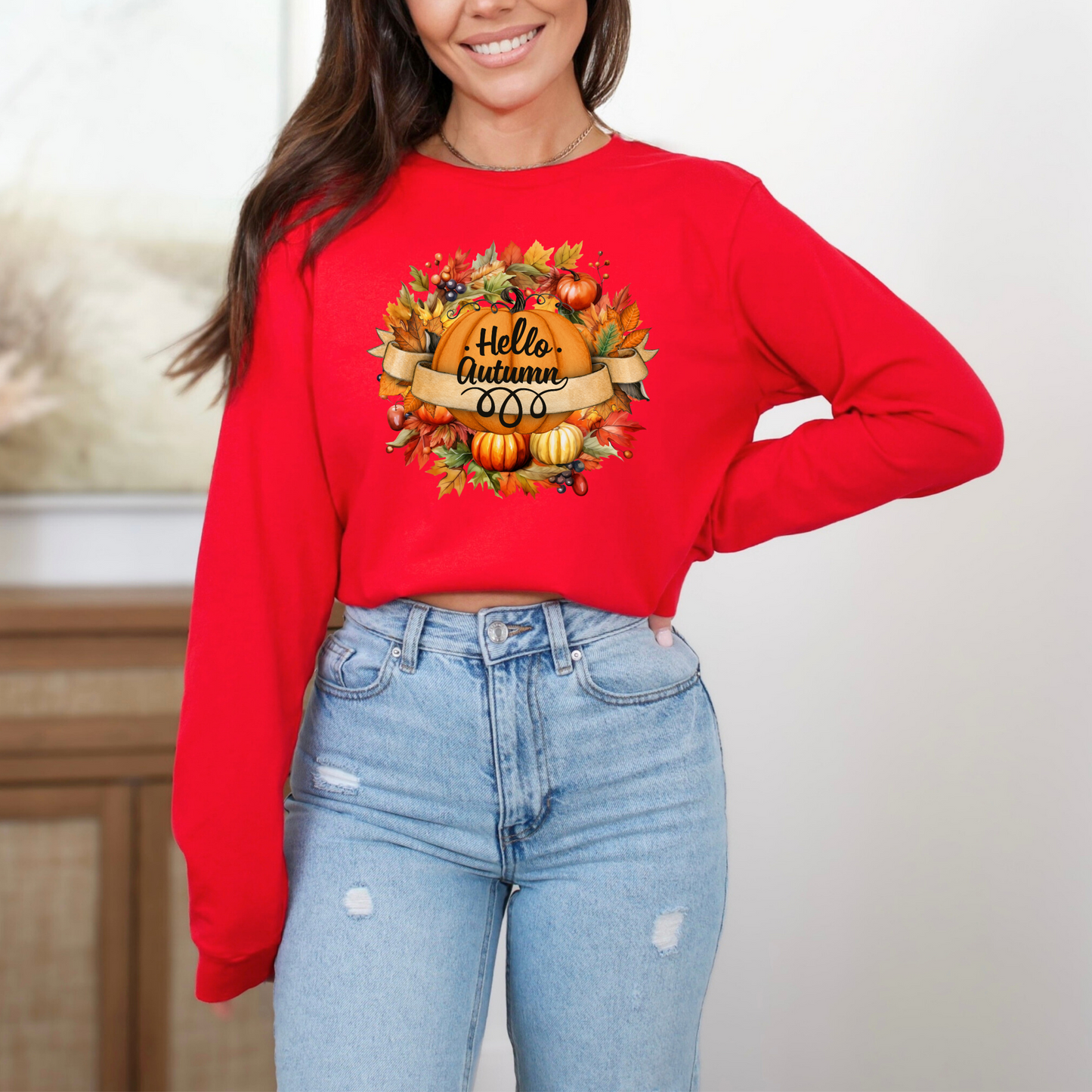 Person wearing a red sweatshirt with 'Hello Autumn' design and blue jeans. featuring a graphic design with the text "Hello Autumn" in black, stylized cursive font on a curved banner. The banner is surrounded by a circular arrangement of colorful autumn leaves, berries, and pumpkins.