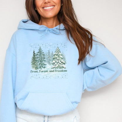 Person wearing a light blue hoodie with a forest design and text on a plain background Light blue hooded sweatshirt featuring a graphic design of a snow-covered forest. The design shows several watercolor-style pine trees in various shades of green and blue, with snow falling around them. Below the trees is the phrase "Frost, Forest, and Freedom" in a matching green font. The entire design is framed by small, delicate snowflake illustrations