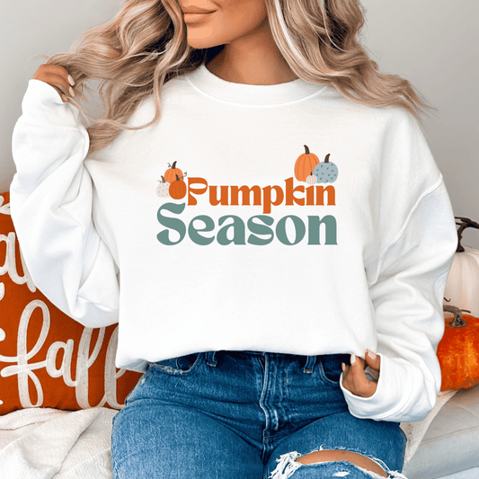 Person wearing a white sweatshirt with 'Pumpkin Season' text and pumpkins graphics sweatshirt featuring a graphic design with the text "Pumpkin Season" in a retro font. The word "Pumpkin" is in orange, and "Season" is in a muted teal color. The design is decorated with a variety of pumpkins in shades of orange, white, and teal, with a small pumpkin vine