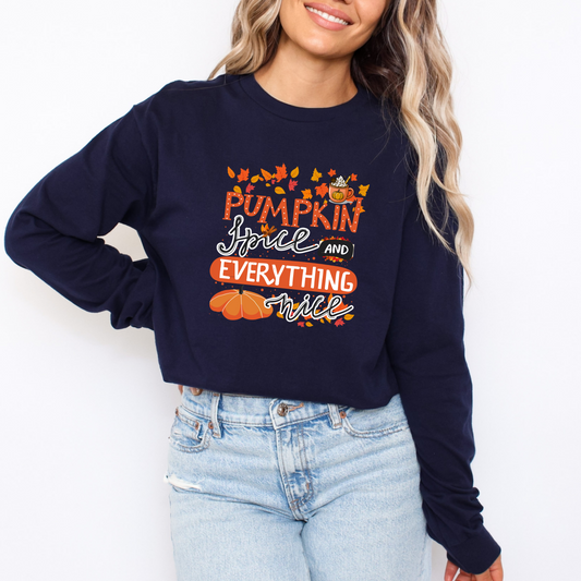 Person wearing a navy sweatshirt with 'Pumpkin Spice and Everything Nice' text and pumpkins design  featuring a colorful graphic design with the text "PUMPKIN" in large orange dotted letters at the top. Below it, "Spice" is written in a black flowing cursive, accented with cinnamon sticks and star anise. To the right, "AND" is in a black banner shape. Underneath, "EVERYTHING" is in stacked orange sans-serif capital letters, and "nice" is written in a black flowing cursive at the bottom. The design is decora