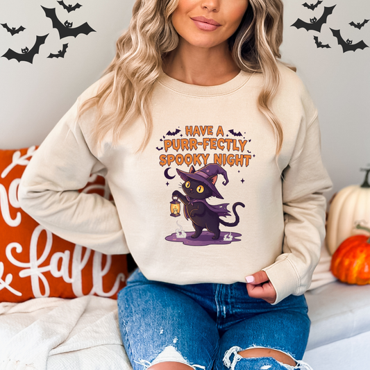 Woman wearing a Halloween-themed sweatshirt with A cartoon-style illustration of a black cat wearing a purple witch hat and cape. The cat is holding a glowing lantern. Above the cat, the text reads, "Have a Purr-fectly Spooky Night." The design also includes bats, a crescent moon, and stars. sitting on a couch with decorative pumpkins and bats in the background.