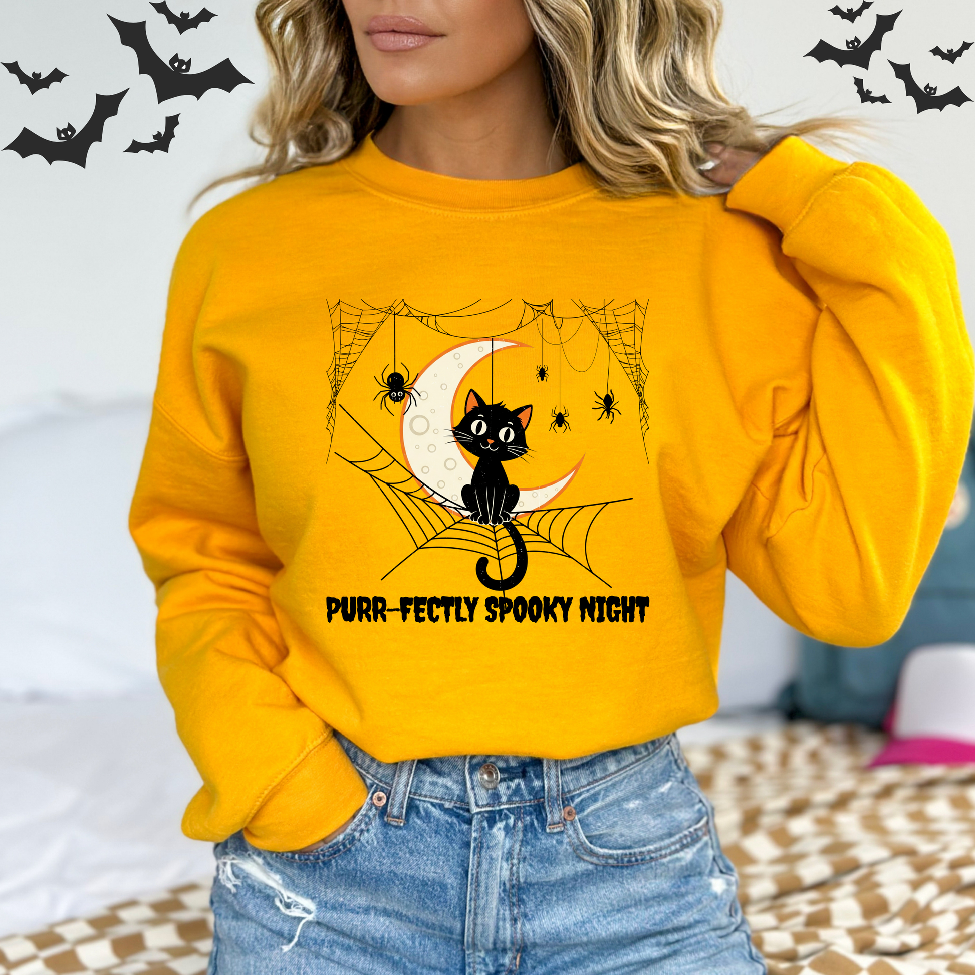 Person wearing a yellow sweatshirt with a Halloween design featuring A vintage-style illustration of a cute black cat sitting on a spiderweb. A large crescent moon is in the background. A small spider is on the web next to the cat. The words "Pur-fectly Spooky Night" are written in a retro font.