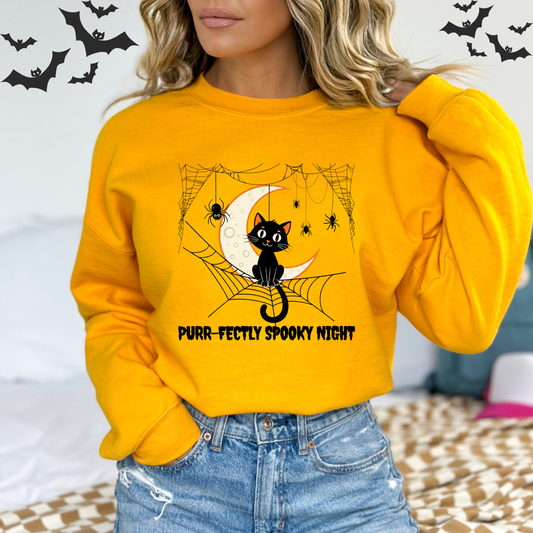 Person wearing a yellow sweatshirt with a Halloween design featuring A vintage-style illustration of a cute black cat sitting on a spiderweb. A large crescent moon is in the background. A small spider is on the web next to the cat. The words "Pur-fectly Spooky Night" are written in a retro font.