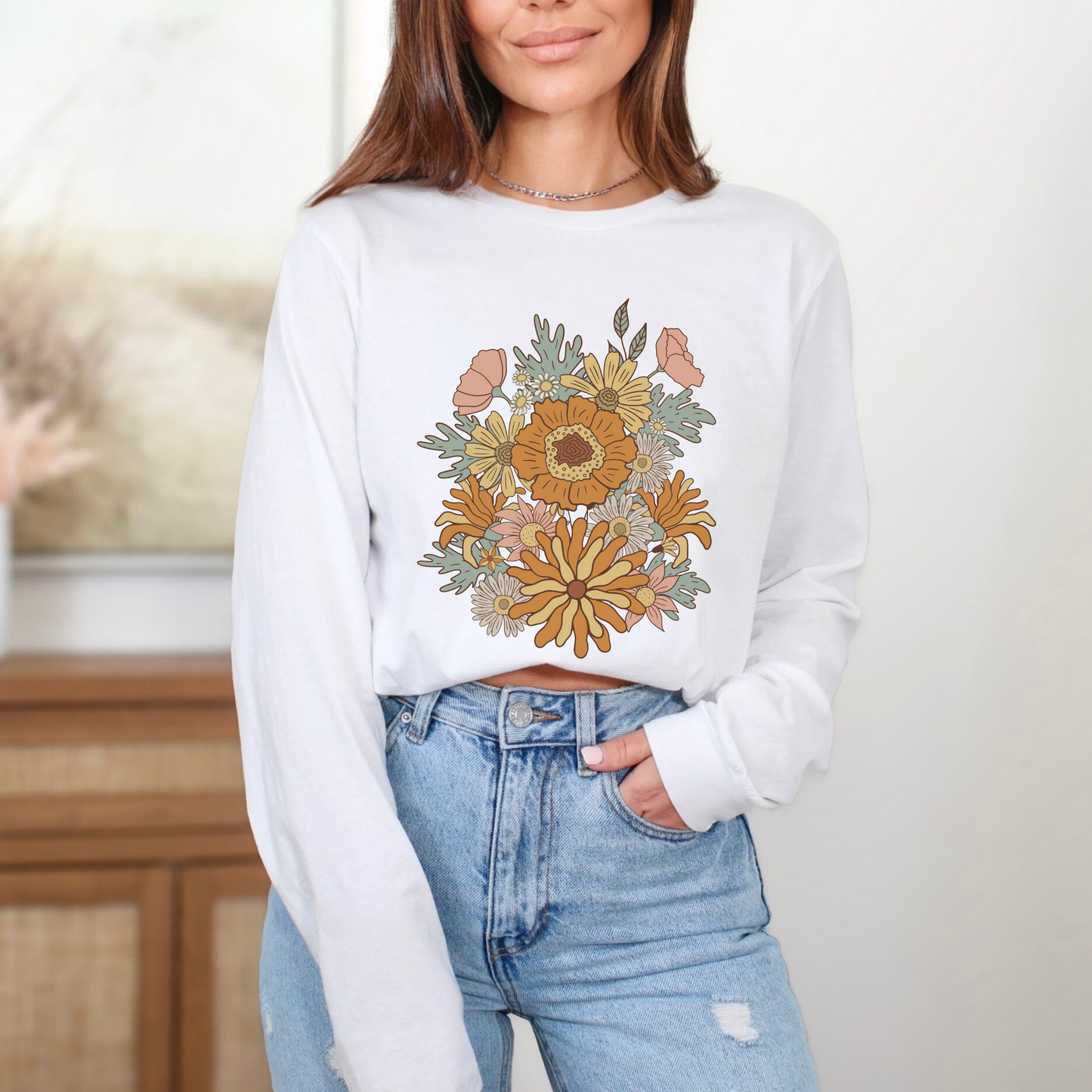 Person wearing a white sweatshirt with a floral design and blue jeans indoors. long sleeve shirt featuring a graphic design of a bouquet of retro-style flowers. The flowers are in shades of orange, yellow, and muted pink with light blue-green leaves and a black outline. The style is reminiscent of a 1970s or groovy aesthetic