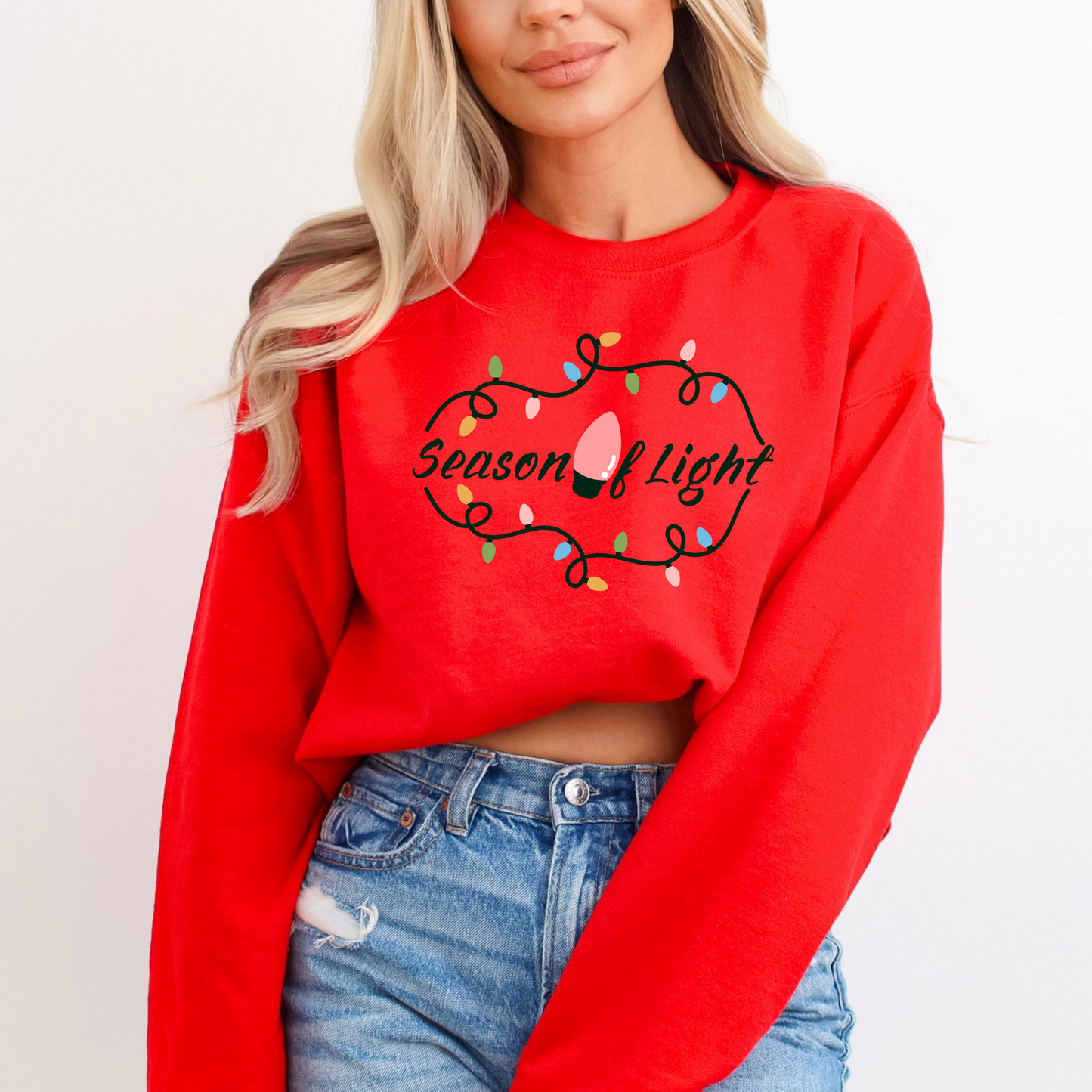 Person wearing a red sweatshirt with 'Season of Light' text and Christmas lights design featuring a graphic design with the word "Autumn" in a stylized cursive font on a curved banner. The banner is wrapped around a large, orange pumpkin. The pumpkin is at the center of a circular arrangement of various fall elements including smaller pumpkins in green and orange, yellow flowers, corn, bundles of wheat, berries, and two lit candles.