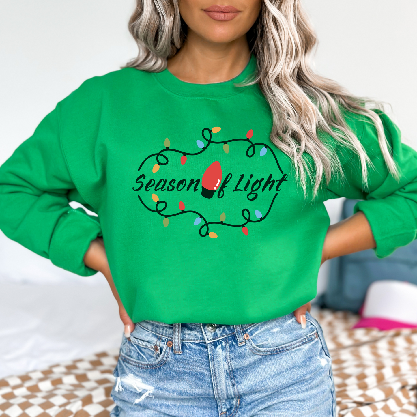 Person wearing a green sweatshirt with 'Season of Light' text and Christmas lights design  featuring a graphic design with the words "Season of Light" in a dark green cursive font. The text is framed by a curving string of colorful holiday lights with a large, bright red light bulb in the center of the phrase. The lights are in shades of red, green, blue, and yellow.