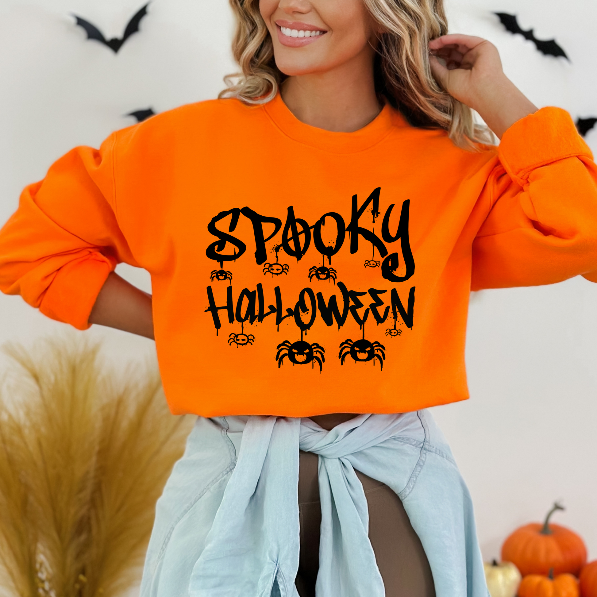 Person wearing an orange sweatshirt , A bold, graffiti-style text illustration that reads "SPOOKY HALLOWEEN" in a dripping font, with various spiders hanging around and crawling on the letters. The design is predominantly black with a distressed or splattered effect.