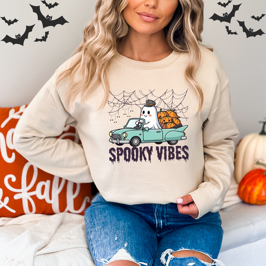 Person wearing a beige sweatshirt withA retro-style illustration of a friendly white ghost wearing a driver's cap, driving a light blue vintage convertible car filled with carved pumpkins. Spiderwebs and dangling skull decorations are in the background, and the text "SPOOKY VIBES" in a dripping, grunge-style font is below the car.