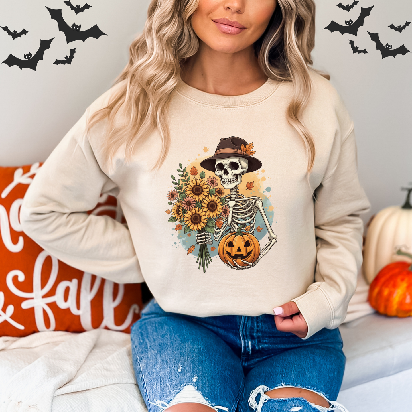 Person wearing a beige sweatshirt with a skeleton holding a pumpkin and flowers, sitting on a couch with Halloween decorations.